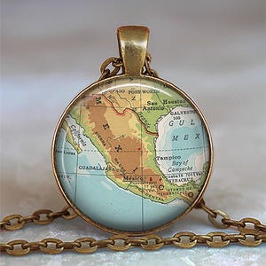 May include: A round pendant necklace with a map of Mexico. The pendant has a gold-colored frame and chain. The map features brown, green, and blue colors, with place names like Mexico City and San Antonio.