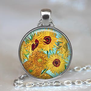 May include: Round silver pendant featuring a painting of sunflowers in yellow, red, and green against a light blue background. The pendant hangs from a silver chain. The artwork is reminiscent of Van Gogh's style.