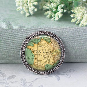 K&ouml;nnte beinhalten: Runde, silberfarbene Brosche mit einer Frankreichkarte. Die Karte ist in Gr&uuml;n-, Gelb- und Beiget&ouml;nen gehalten, mit St&auml;dtenamen. Die Brosche hat einen dekorativen, perlenbesetzten Rand.