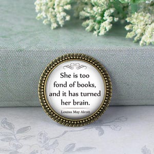 May include: A circular, antique-style pin with a quote by Louisa May Alcott: "She is too fond of books, and it has turned her brain." The pin features a bronze-coloured border and a white central design. A literary-themed accessory.