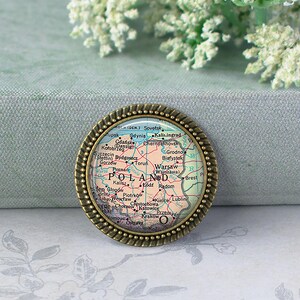 May include: A round, antique-style pin featuring a map of Poland. The map is in shades of beige, green, and blue, with cities labeled. The pin has a bronze-colored, rope-textured border. The word "POLAND" is prominently displayed.