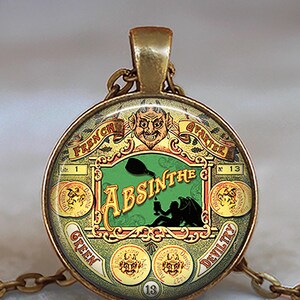 May include: A round, antique-style pendant with a gold-tone chain. The pendant features a green and gold design with the words "ABSINTHE" and "FRENCH QUARTER." The design includes a devil figure and other decorative elements.