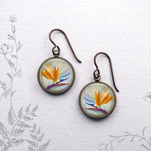 May include: A pair of round earrings with a bronze-colored frame. Each earring features a vibrant image of a bird of paradise flower in shades of orange, yellow, purple, and blue. The earrings have a hook style for pierced ears.