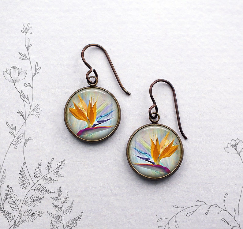 May include: A pair of round earrings with a bronze-colored frame. Each earring features a vibrant image of a bird of paradise flower in shades of orange, yellow, purple, and blue. The earrings have a hook style for pierced ears.