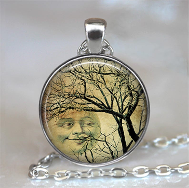 May include: A round silver pendant necklace featuring a vintage-style illustration of a smiling moon face partially obscured by dark, leafless tree branches against a pale, textured background, suspended from a matching silver link chain.