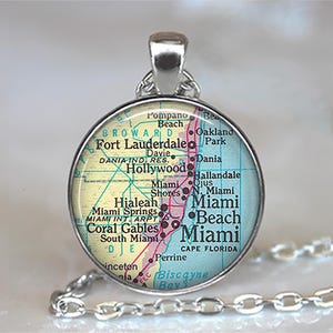 May include: A silver-toned pendant necklace featuring a round charm with a map of Miami, Florida. The map includes city names like Fort Lauderdale, Miami Beach, and Coral Gables. The pendant hangs from a delicate chain.