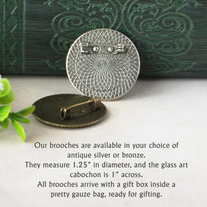 a round, antique-looking silver or bronze brooch with a detailed design, placed on a white surface next to a green plant. The text provides information about the brooches, stating that they are available in silver or bronze and measuring 1.25" in diameter. The glass art cabochon is 1" across, and all brooches come with a gift box inside a pretty gauze bag, ready for gifting.