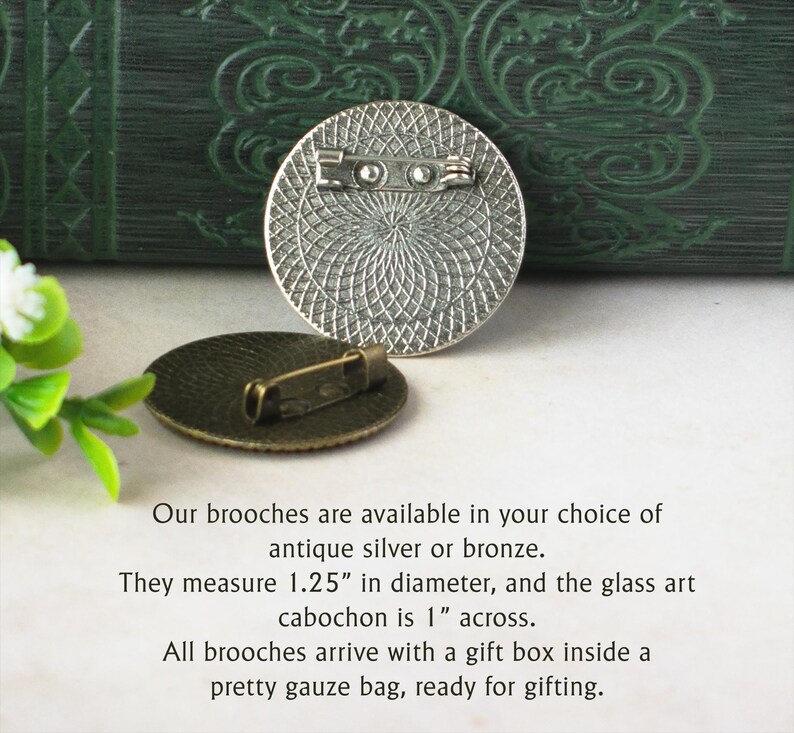 a round, antique-looking silver or bronze brooch with a detailed design, placed on a white surface next to a green plant. The text provides information about the brooches, stating that they are available in silver or bronze and measuring 1.25" in diameter. The glass art cabochon is 1" across, and all brooches come with a gift box inside a pretty gauze bag, ready for gifting.