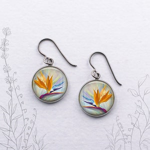 May include: A pair of round, silver-tone earrings. Each earring features a colorful image of a bird of paradise flower with orange, yellow, purple, and blue hues. The earrings hang from curved ear wires.