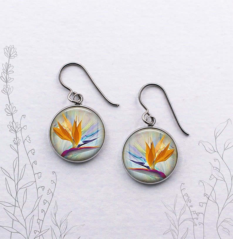 May include: A pair of round, silver-tone earrings. Each earring features a colorful image of a bird of paradise flower with orange, yellow, purple, and blue hues. The earrings hang from curved ear wires.
