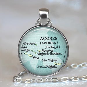 May include: Silver pendant necklace featuring a round charm with a map of the Azores islands. The map is in shades of green and white, with text indicating the islands and their locations. The chain is silver.