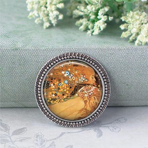 Puede incluir: Un broche redondo plateado que presenta una pintura famosa con tonos c&aacute;lidos de oro, naranja y azul. La obra de arte representa dos figuras abraz&aacute;ndose, rodeadas de detalles florales y abstractos. El broche tiene un borde texturizado en forma de cuerda.