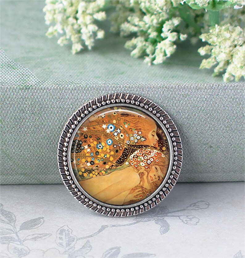 Puede incluir: Un broche redondo plateado que presenta una pintura famosa con tonos c&aacute;lidos de oro, naranja y azul. La obra de arte representa dos figuras abraz&aacute;ndose, rodeadas de detalles florales y abstractos. El broche tiene un borde texturizado en forma de cuerda.