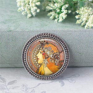 May include: Round silver-toned brooch featuring an Art Nouveau style illustration of a woman with flowing auburn hair and ornate headwear. The brooch has a beaded edge and text around the perimeter, including "SAVONNERIE DE BAGNOLET" and "ALFRED SCHWEIZER".
