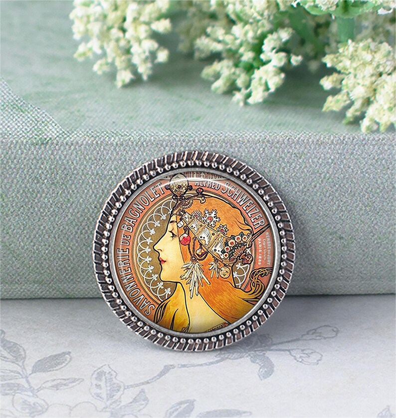 May include: Round silver-toned brooch featuring an Art Nouveau style illustration of a woman with flowing auburn hair and ornate headwear. The brooch has a beaded edge and text around the perimeter, including "SAVONNERIE DE BAGNOLET" and "ALFRED SCHWEIZER".