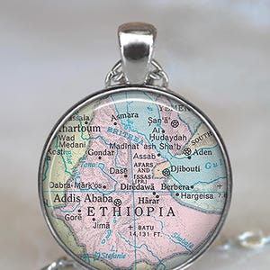 May include: A round silver pendant featuring a map of Ethiopia and surrounding areas. The map is in shades of pink, green, and blue, with place names in black. The pendant hangs from a silver chain.