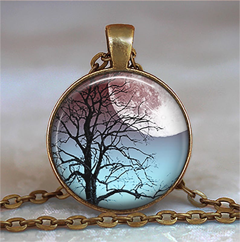 K&ouml;nnte beinhalten: Eine bronzene Halskette mit einem runden Glasanh&auml;nger, der eine Silhouette eines Baumes vor einem rosa und blauen Mond zeigt.