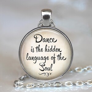 May include: A round silver pendant necklace featuring a glass cabochon with a cream-colored background and black cursive text that reads: Dance is the hidden language of the Soul, complete with a decorative flourish at the bottom of the quote.