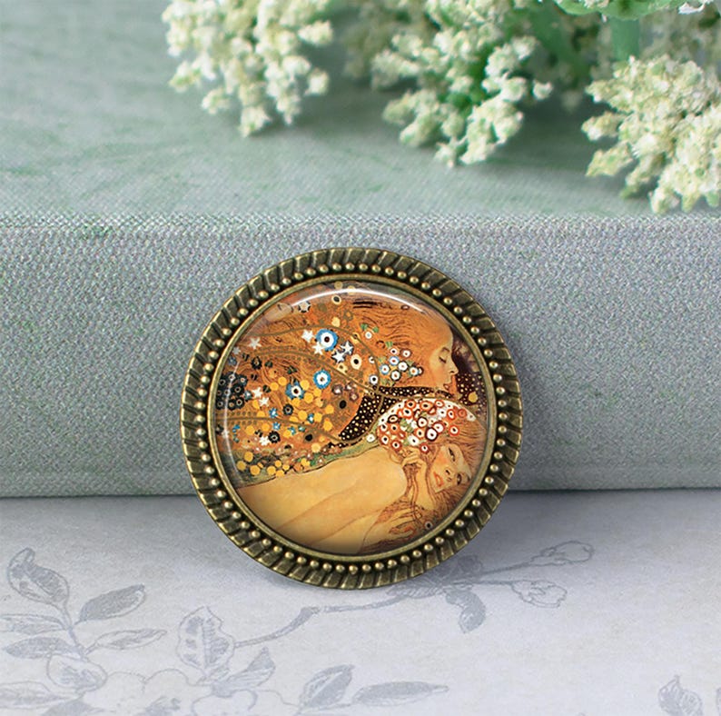 Puede incluir: Broche redondo en tonos bronce con una imagen detallada de una pintura con figuras en tonos c&aacute;lidos de oro, naranja y marr&oacute;n. La obra de arte est&aacute; enmarcada por un borde texturizado. El broche est&aacute; sobre un fondo suave y neutro.