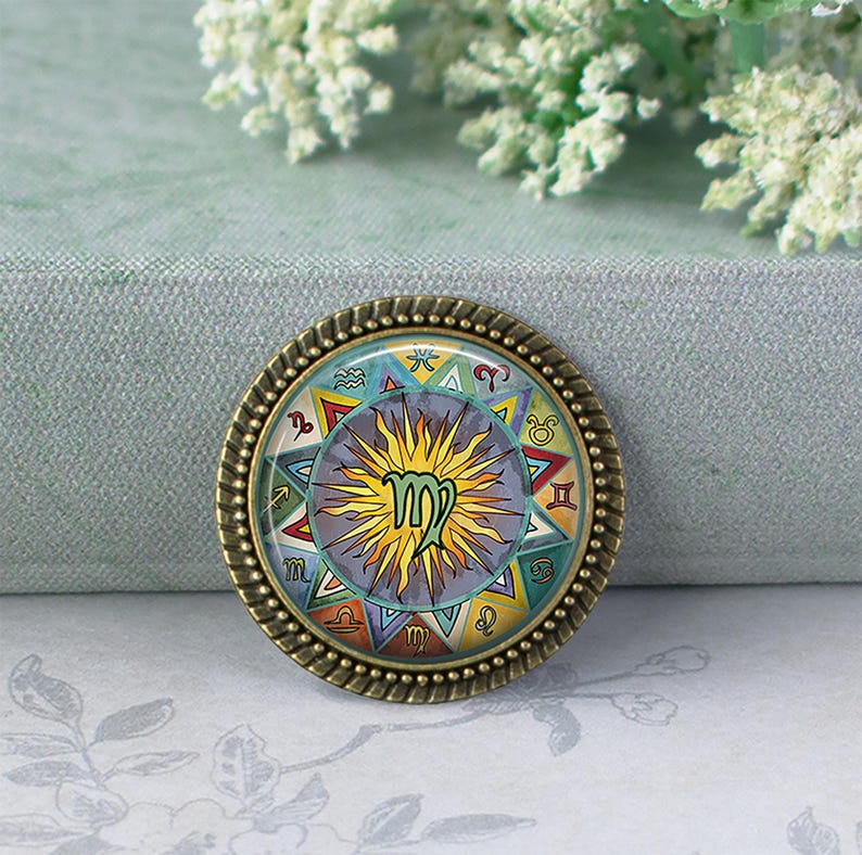May include: A round, bronze-colored Virgo zodiac sign pendant. The pendant features a colorful design with the Virgo symbol at the center, surrounded by other zodiac symbols and a sunburst pattern. The pendant has a detailed, textured border.