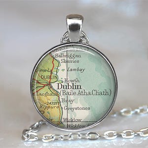 May include: A silver-toned pendant necklace featuring a circular charm with a map of Dublin, Ireland. The map shows the city and surrounding areas in green, yellow, and red. The pendant hangs from a silver chain.