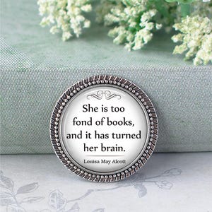 May include: A circular silver-coloured pin featuring a quote by Louisa May Alcott: "She is too fond of books, and it has turned her brain." The pin has a decorative border and a white background.