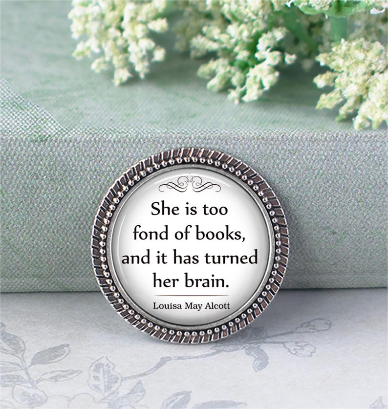 May include: A circular silver-coloured pin featuring a quote by Louisa May Alcott: "She is too fond of books, and it has turned her brain." The pin has a decorative border and a white background.