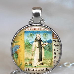 May include: A silver pendant necklace with a round glass charm featuring a vintage illustration of a man in a brown robe holding a book and a shovel. The background is a yellow and green landscape with blue flowers. The text on the charm reads "De famur lade is facrus pecabur".