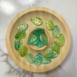 May include: A wooden bowl holds several leaf-shaped beads in shades of green and gold. The beads are arranged in a circular pattern, with some inside a smaller, central bowl. The leaves have detailed vein patterns.