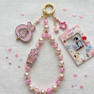 May include: A beaded keychain featuring a gold ring, alternating pink translucent and opaque white beads, and three charms: a pink character in a circle, a pink boba tea cup, and a rectangular BT21 photo frame containing a portrait of a person.