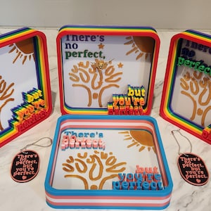 Op de afbeelding: Een verzameling vierkante schaduwboksen met regenboogkleurige frames en de tekst "There's no perfect, but you're perfect". Elke doos heeft een boomontwerp en een zon. Er zijn ook bijpassende sleutelhangers.