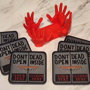 May include: A set of square coasters in grey and black, featuring the text "DONT DEAD OPEN INSIDE" and space for custom text. A red hand-shaped sculpture is also visible. These coasters are likely for home use.