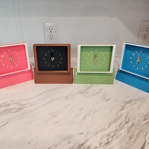May include: Four analogue clocks shaped like vintage desktop computers, each featuring a screen-style clock face with numbers one through twelve and gold-coloured hands. The clocks are coloured pink, brown, light green, and blue, with integrated keyboards.