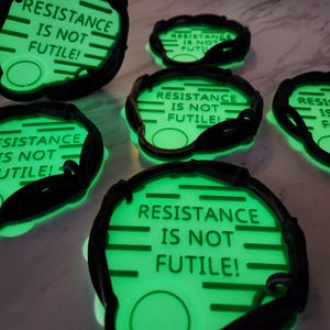 May include: Several glowing green coasters with the text "RESISTANCE IS NOT FUTILE!" in white. The coasters have a black border and a circular design. The image is taken from a slightly elevated angle.