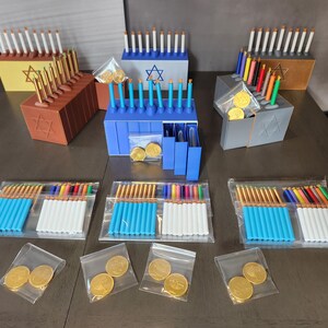 May include: A selection of Hanukkah menorahs in shades of brown, yellow, blue, and silver. Each menorah holds candles and is presented with small bags of gold coins. The menorahs are arranged on a dark wooden surface.