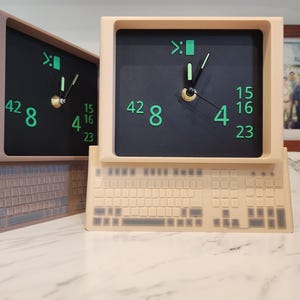 May include: Two retro-style computer clocks with a beige frame and keyboard base. The clock face is black with green numerals and hands. The time is displayed as 8:16. A framed photo is in the background.