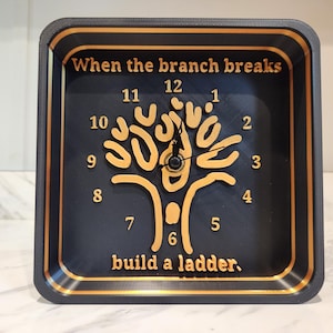 Może przedstawiać: Kwadratowy zegar z czarną tarczą i złotymi akcentami. Zegar posiada motyw drzewa i cytat: „When the branch breaks build a ladder”. Wskazówki zegara są czarne. Cyfry są złote.