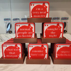 May include: A stack of square coasters in brown holders, each featuring a white background with a red blood splatter graphic and text. The text reads: i'm a very neat monster, normal people are so hostile, tonight's the night, what was that morgan?, remember harry's code, and surprise motherfucker.