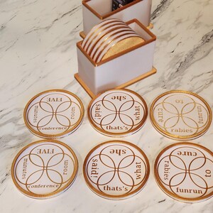 May include: A collection of six white and gold coasters, each featuring a different phrase, such as "FIVE minutes conference room" and "what she said!". The coasters have a circular shape, gold edging, and a gold floral pattern. A matching holder containing coasters is also present.