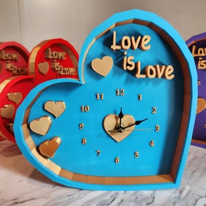 May include: Heart-shaped wall clocks in various colors, including blue, red, pink, and purple. Each clock features the phrase "Love is Love" and heart-shaped accents. The clock hands are black, and the numbers are visible.