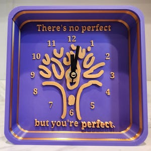 Pode incluir: Um relógio quadrado roxo com acabamento dourado e um design dourado em forma de árvore no centro. O mostrador do relógio tem as palavras "There's no perfect" no topo e "but you're perfect" na parte inferior.
