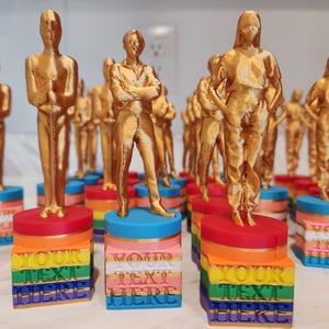 May include: A collection of gold-colored statuettes on rainbow-colored bases. Each base has the text "YOUR TEXT HERE" in a stacked, colorful design. The statuettes depict figures in various poses, suggesting awards or trophies.