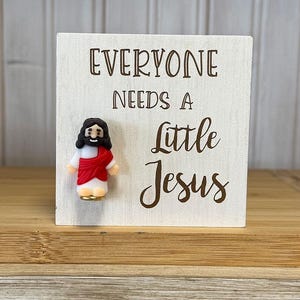 May include: White wooden sign with the text "Everyone needs a little Jesus" and a small figurine of Jesus in red and white.