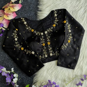 May include: Black long-sleeved blouse with white and yellow floral embroidery. The embroidery is in a vine pattern along the neckline, front, and sleeves. The blouse is made of a shiny, satin-like fabric.