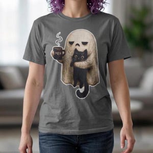 Peut inclure: Un t-shirt gris anthracite avec un motif représentant un fantôme beige tenant un chat noir et une tasse sombre sur laquelle est écrit MEH. Le fantôme a une expression neutre et de la vapeur s'échappe de la tasse en un trait blanc.