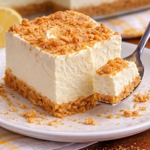May include: A square slice of cheesecake with a crumbly topping and crust, served on a white plate. A small piece of the cheesecake is on a spoon. The dessert has a creamy, off-white filling and a golden-brown crust.