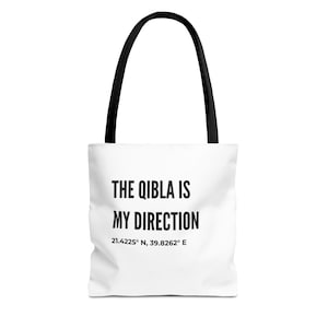 May include: White tote bag with black handles featuring the text "THE QIBLA IS MY DIRECTION" in black. Below the text are the coordinates 21.4225° N, 39.8262° E. The bag is designed for carrying items.