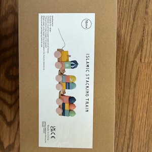 May include: A wooden Islamic stacking train toy with colorful blocks and wheels. The box is brown with the text "ISLAMIC STACKING TRAIN" and "18m+". The toy is designed for children 18 months and older.