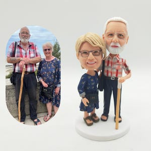 Könnte beinhalten: Eine personalisierte Wackelkopf-Figur eines Paares, das auf einem weißen Sockel steht. Die männliche Figur trägt ein rot-weiß kariertes Hemd und eine schwarze Hose und hält einen Stock. Die weibliche Figur trägt ein blaues Kleid. Ein Foto des Paares ist im Hintergrund.