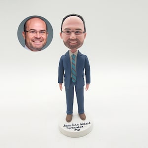 May include: A personalised bobblehead figure of a man in a blue suit, brown shoes, and a striped tie. The figurine stands on a white base with the name "Juan Jose Gilbert Fernandez. MD". A photo of the man is in the upper left corner.